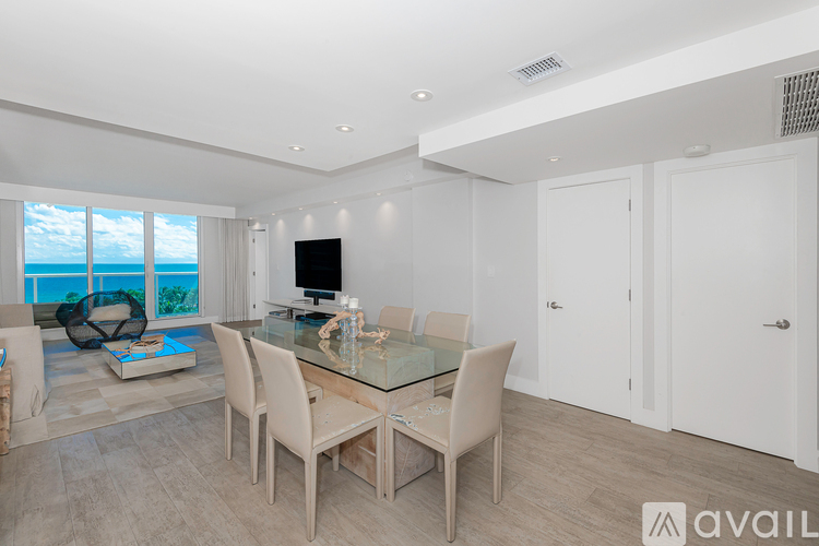 A modern dining room with a glass table and chairs, a television, and a view of the ocean through the windows.