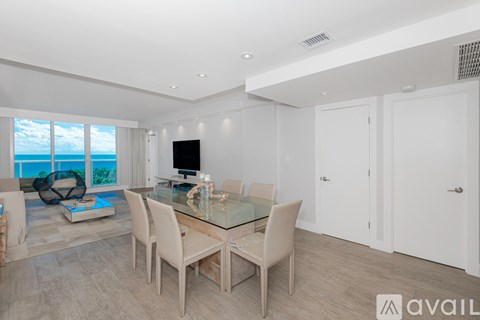 A modern dining room with a glass table and chairs, a television, and a view of the ocean through the windows.