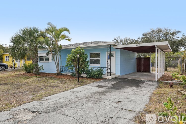 A blue house with a white door and windows is surrounded by trees and has a cracked driveway.