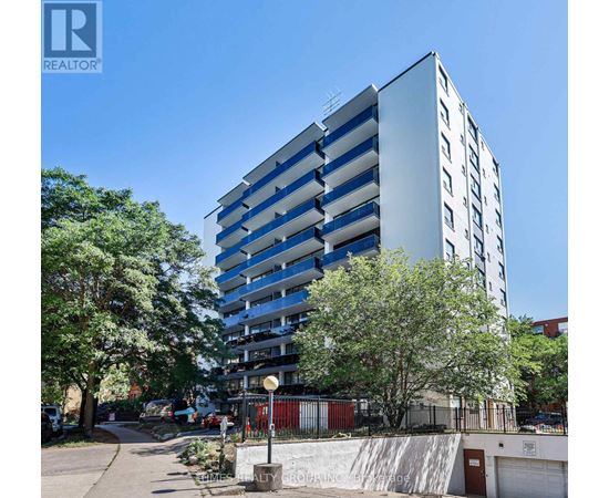 A tall building with a blue and white facade is surrounded by trees and a clear sky.