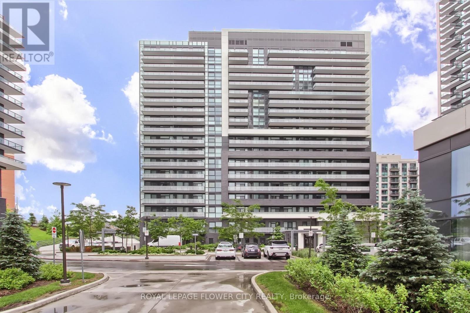 A tall building with a grey facade is surrounded by greenery and other buildings.