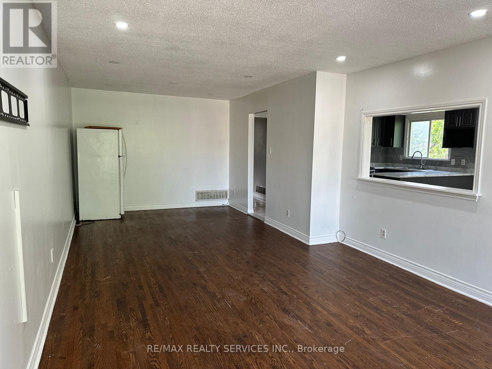 A spacious room with wooden floors and white walls, featuring a window with a view of a green outdoor area.