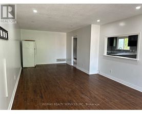 A spacious room with wooden floors and white walls, featuring a window with a view of a green outdoor area.
