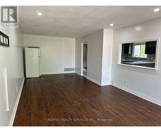 A spacious room with wooden floors and white walls, featuring a window with a view of a green outdoor area.