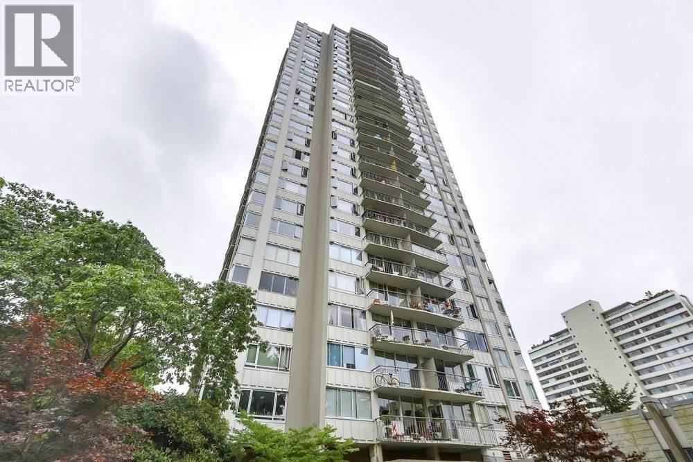 A tall residential building with balconies and windows is surrounded by green trees.