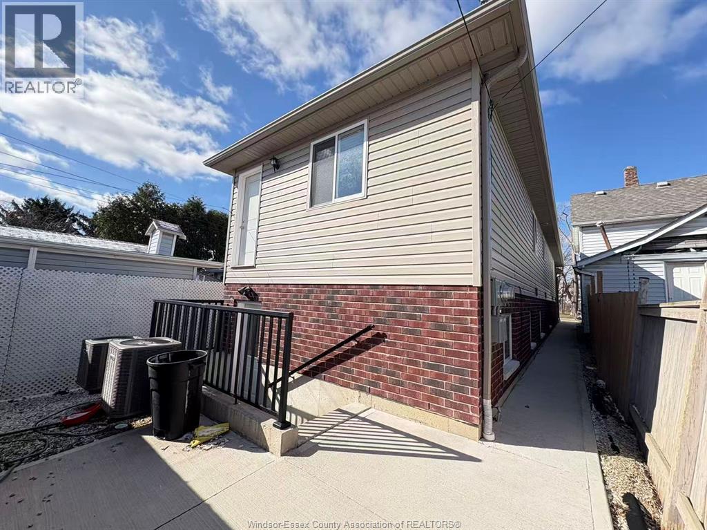A house with a beige siding and a brick wall is shown with a black trash can and a black railing.