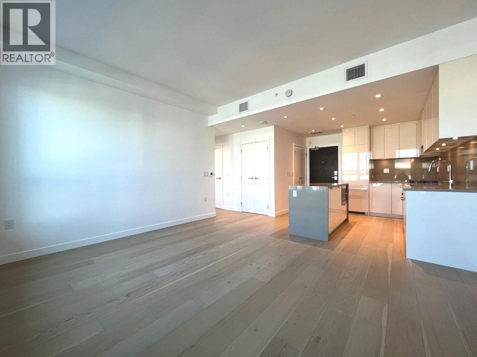 A spacious room with wooden flooring and a kitchen area in the background.