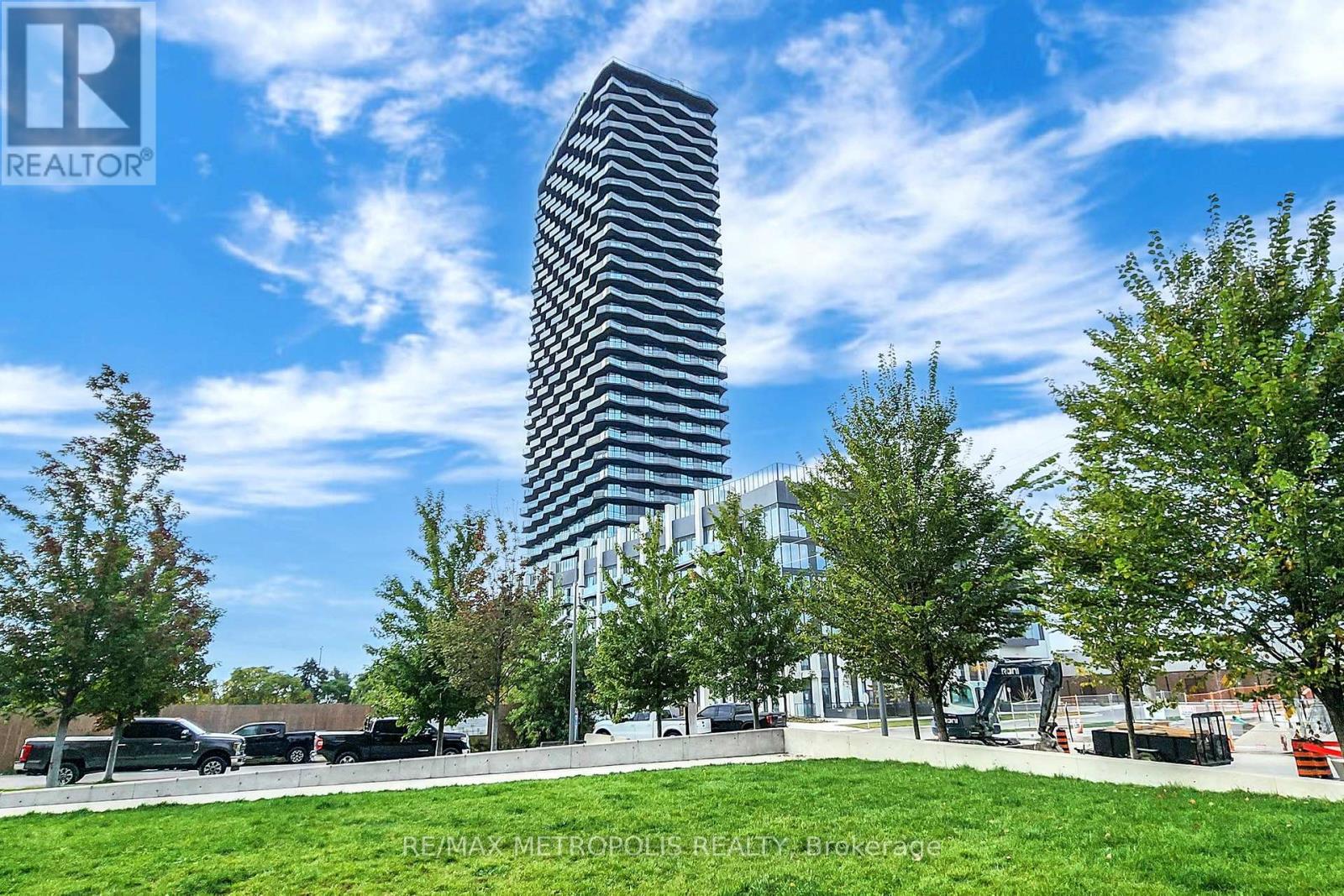 A tall building with a striped facade stands in a grassy area with trees and a parking lot in the foreground.