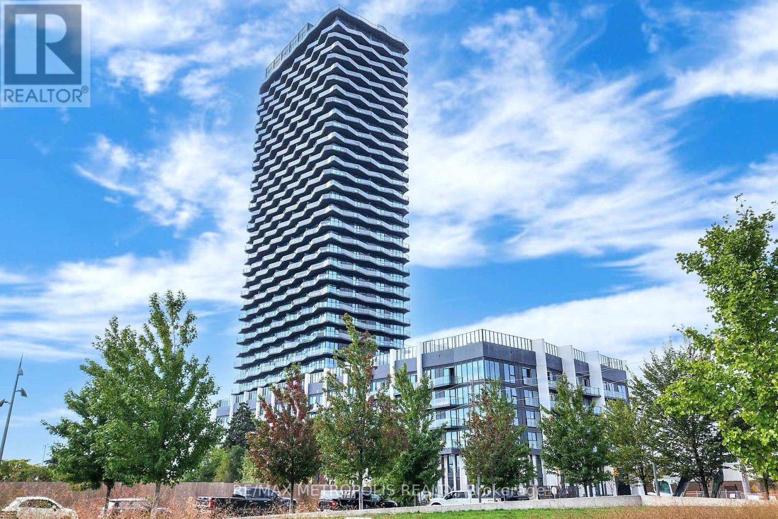 A tall building with a glass facade stands in front of a parking lot and other buildings.