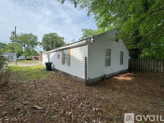 A small white house with a brown roof is surrounded by trees and has a trash can in front of it.