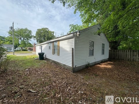 A small white house with a brown roof is surrounded by trees and has a trash can in front of it.