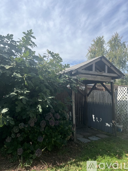 A small wooden structure with a triangular roof is surrounded by greenery and a wooden fence.