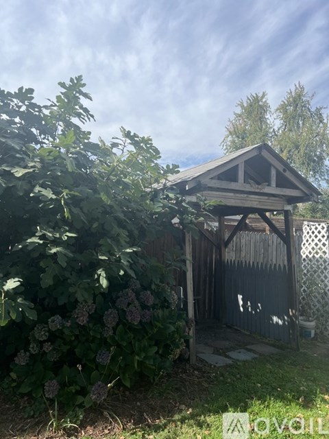 A small wooden structure with a triangular roof is surrounded by greenery and a wooden fence.