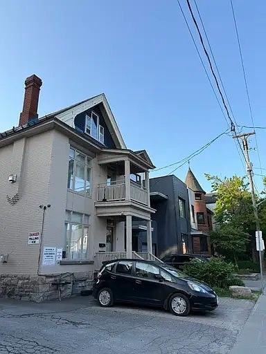 A black car is parked in front of a house with a for sale sign.