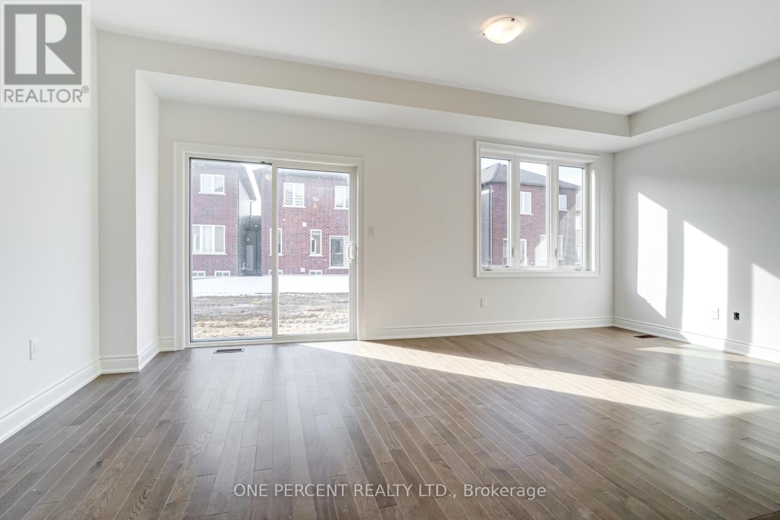 A room with wooden floors and white walls, with a view of a red brick building outside through the windows.