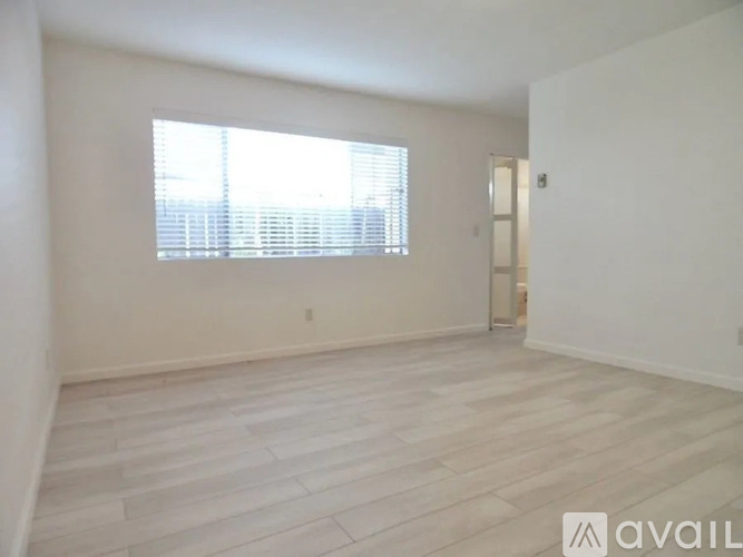 A spacious room with light wood flooring and a large window with blinds.