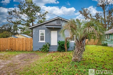 A house with a blue exterior and a white door is surrounded by a wooden fence and a palm tree.