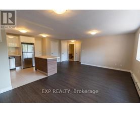 A spacious room with wooden flooring and white walls, with a kitchen area visible in the background.