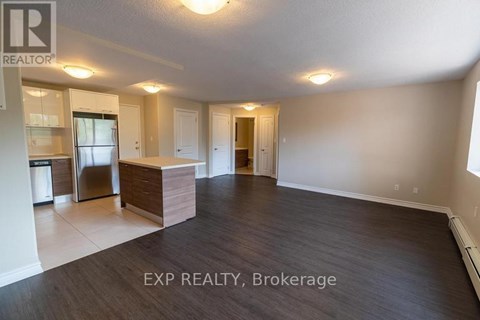 A spacious room with wooden flooring and white walls, with a kitchen area visible in the background.