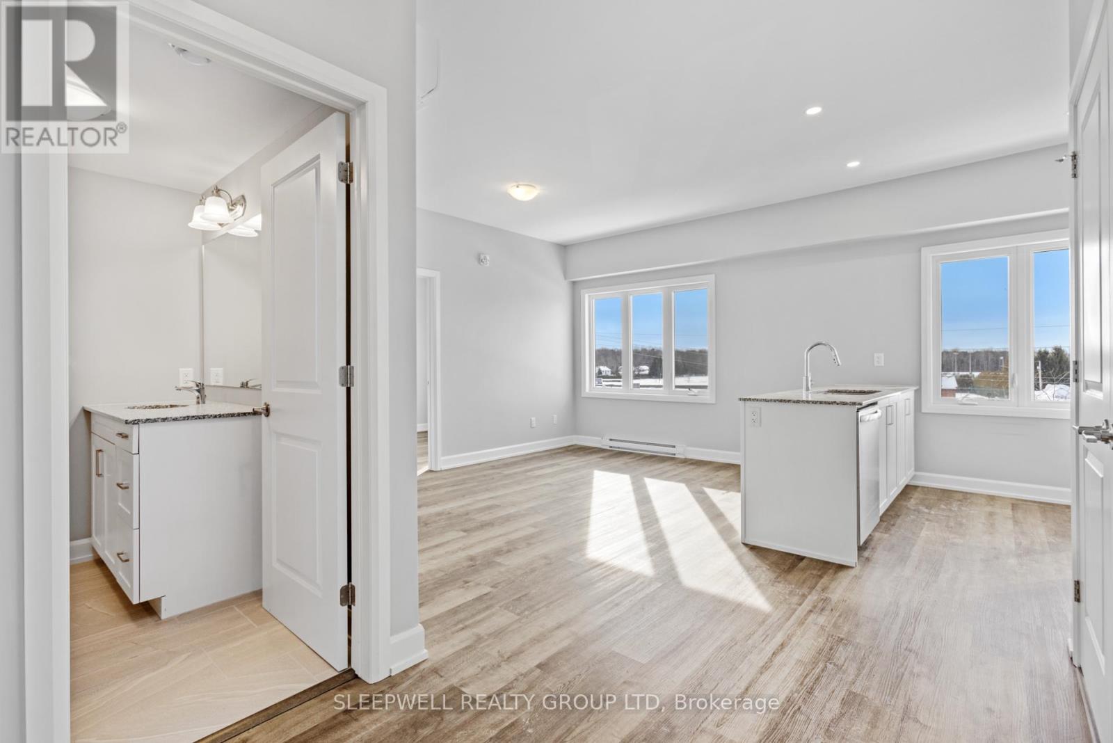 A realtor photo of a white kitchen with wood floors and a view of the outdoors through the windows.