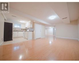 A spacious room with wooden flooring and white walls, with a kitchen area visible in the background.