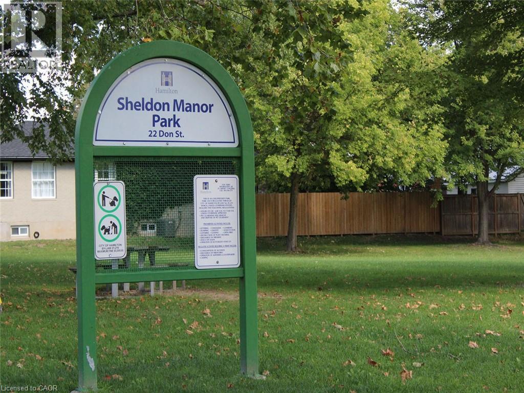 A green sign for Sheldon Manor Park is in front of a fence and trees.