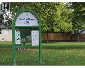 A green sign for Sheldon Manor Park is in front of a fence and trees.