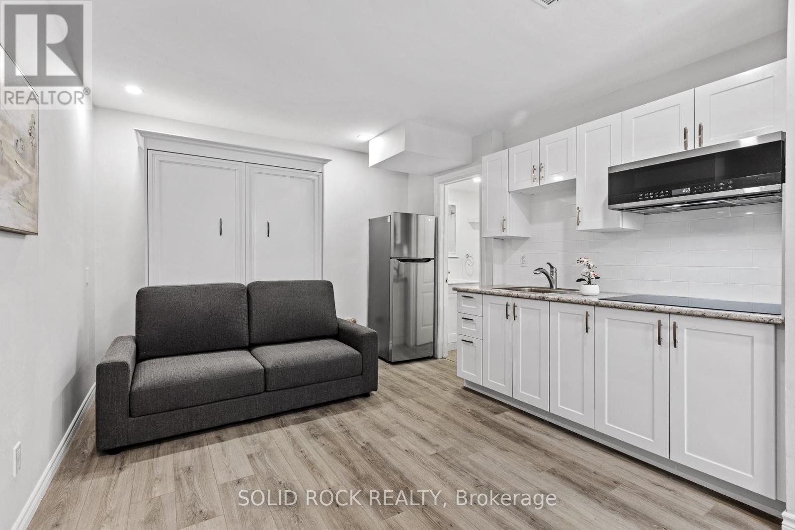 A living room with a grey couch and a kitchen area with white cabinets.