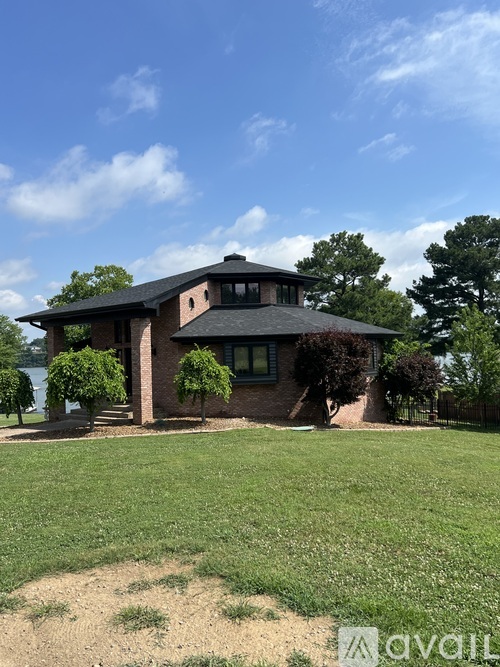 A house with a black roof and a chimney is surrounded by a grassy lawn and trees.
