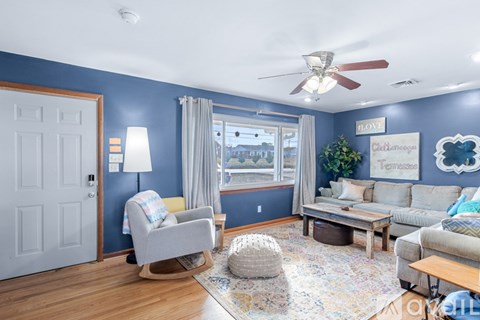 A living room with a white door, a grey sofa, a wooden coffee table, a rug, a ceiling fan, and a wall with a sign that says "CHRISTMAS".