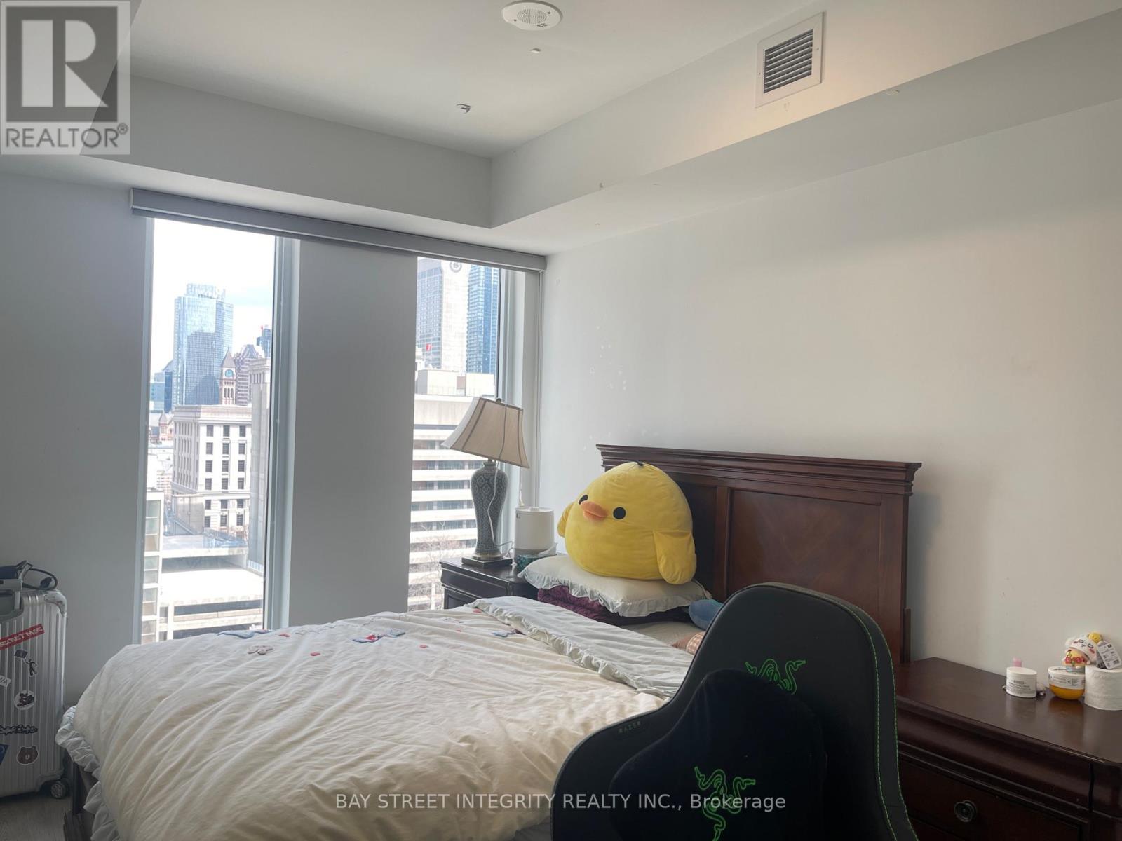 A bedroom with a bed, a yellow duck pillow, and a window overlooking a cityscape.