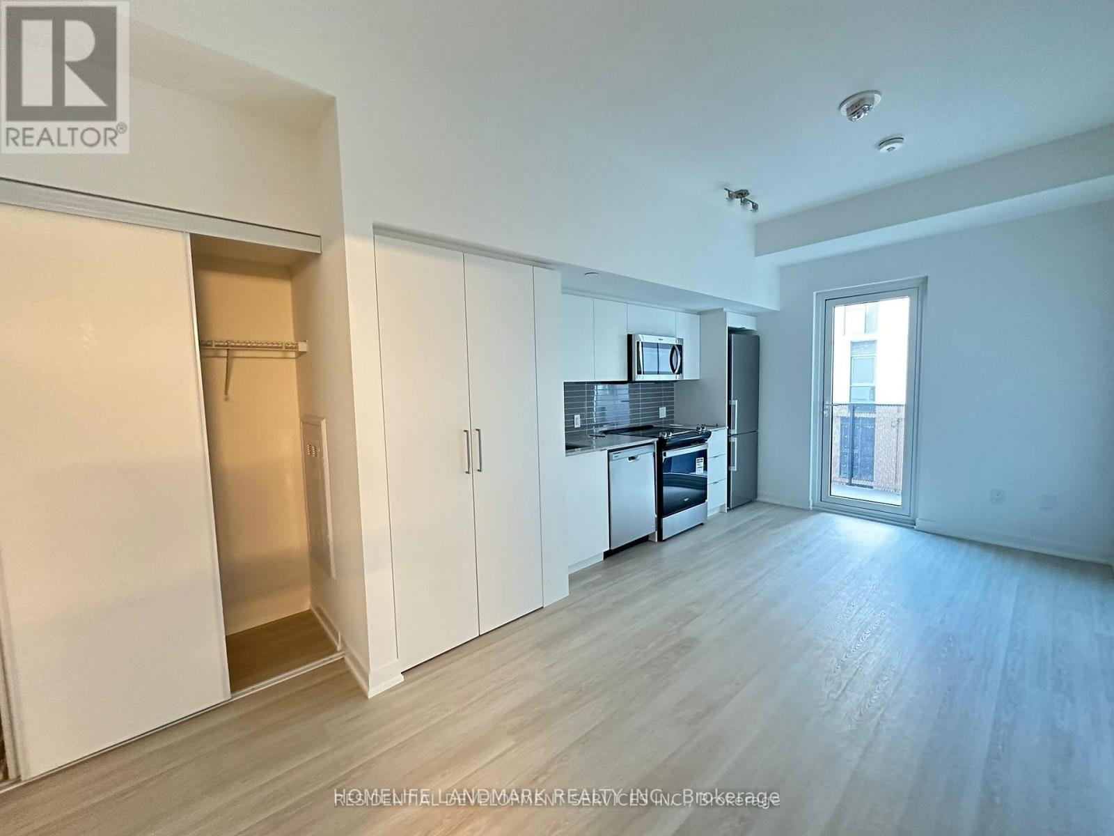 A spacious, empty room with white walls and wooden flooring, featuring a kitchenette with a refrigerator, microwave, and oven.