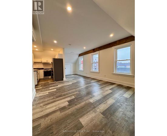 A spacious room with wood flooring and a kitchen area with a refrigerator.