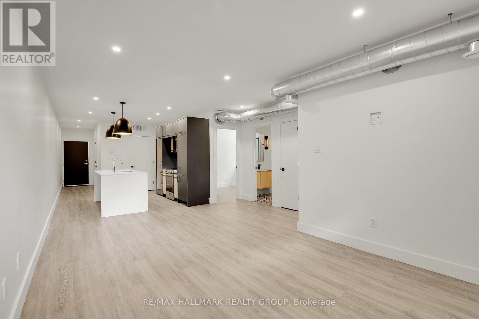 A spacious room with white walls and wooden flooring, featuring a kitchenette with a sink and a refrigerator.