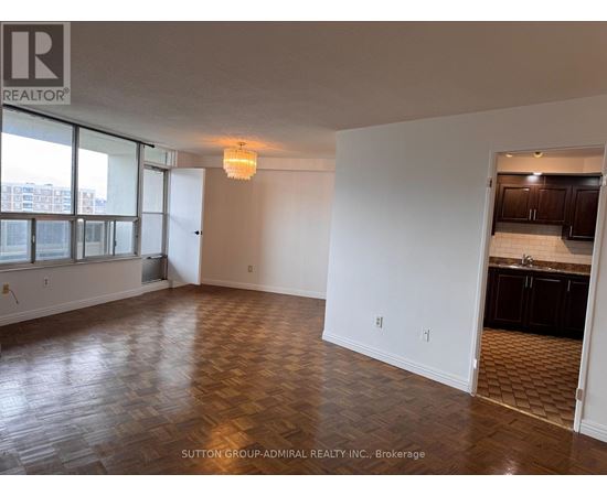 A room with wooden flooring and a kitchen area in the background.