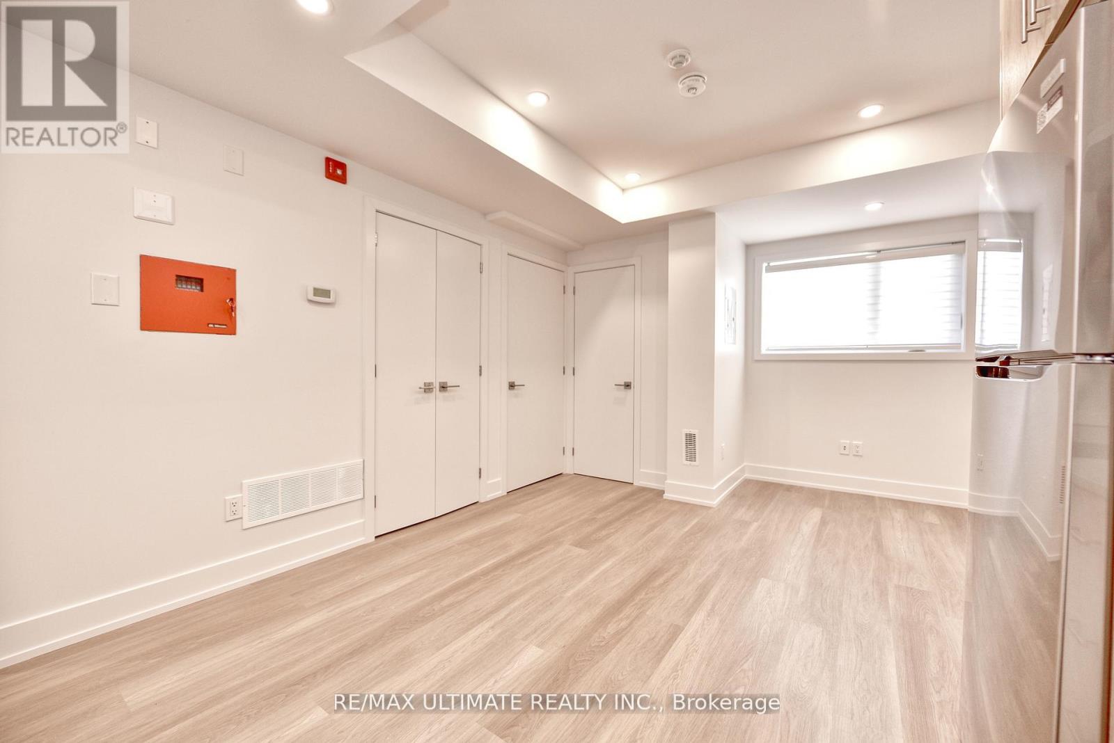A spacious room with white walls and wooden flooring, featuring a row of white cupboards and a window with blinds.