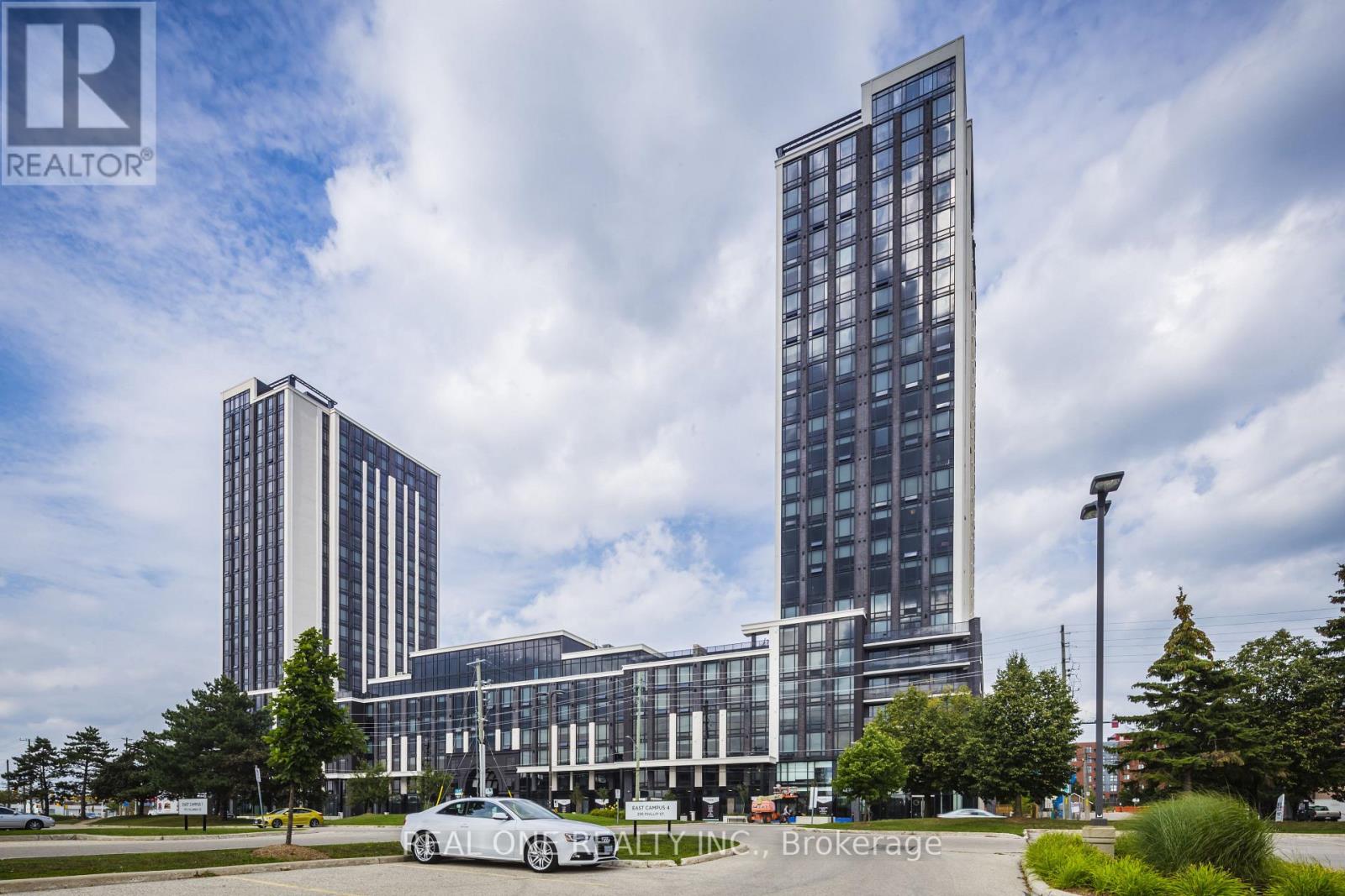 A tall building with a glass facade stands in front of a smaller building and trees.