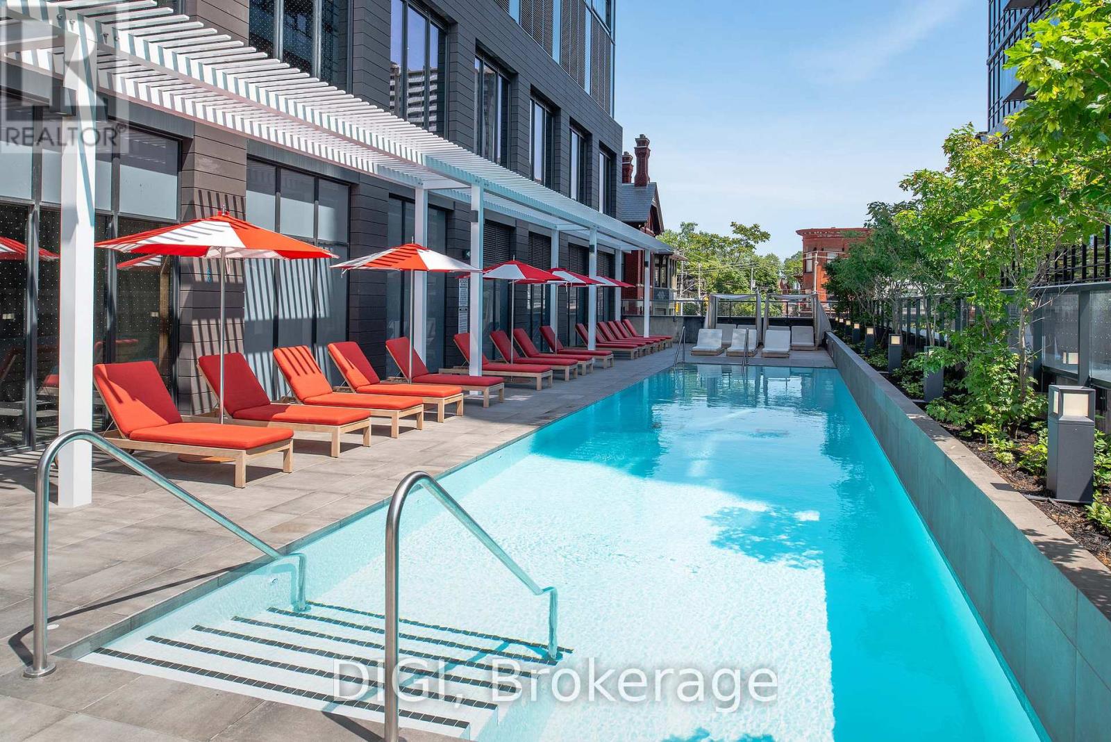 A pool with red chairs and umbrellas is surrounded by buildings.