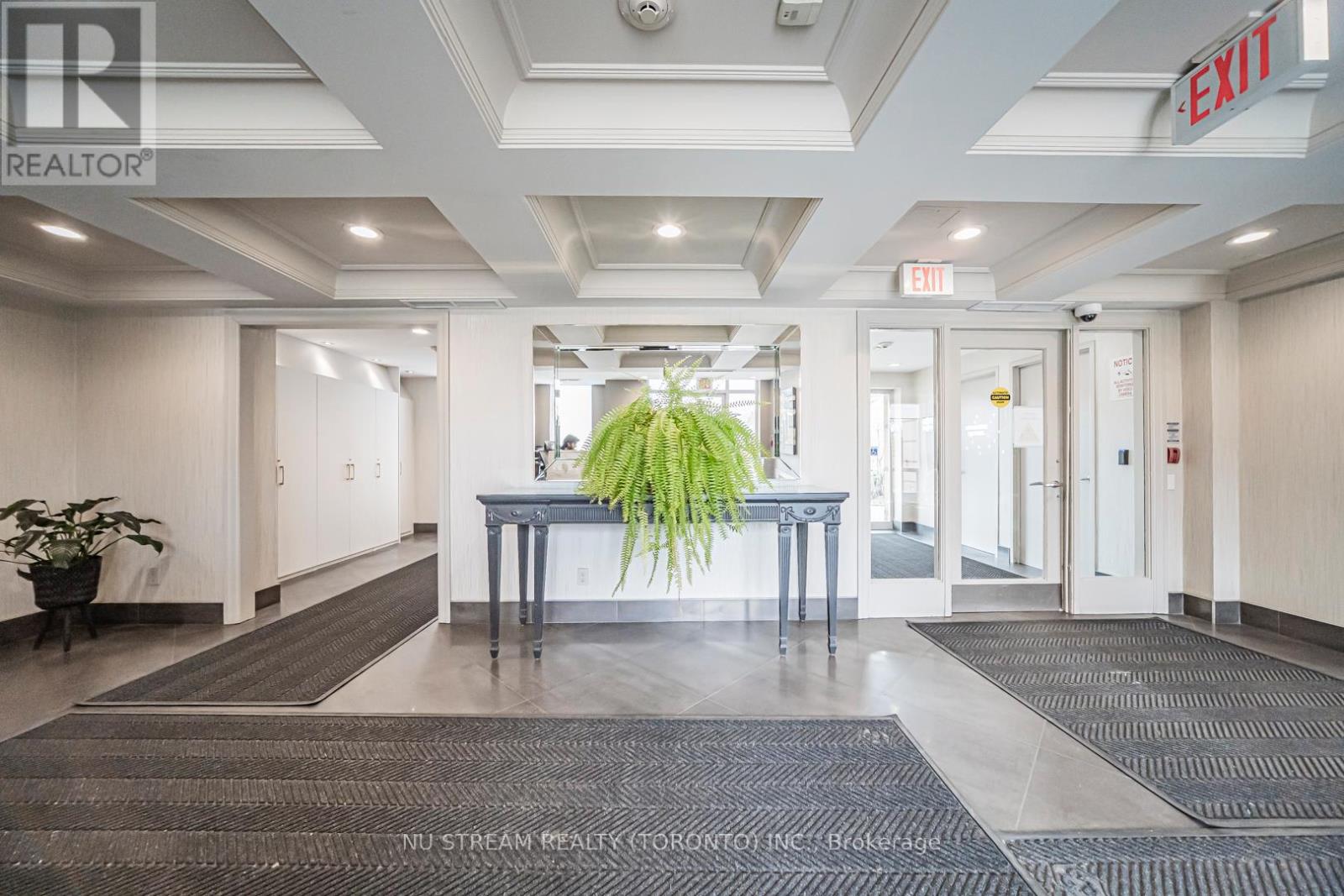 A hallway with a plant in the middle and a red exit sign above.