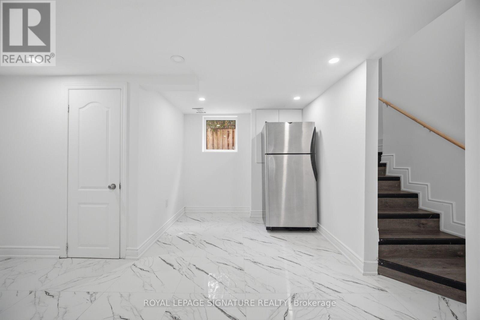 A spacious, white-walled room with a marble floor, a stainless steel refrigerator, and a staircase with wooden steps.