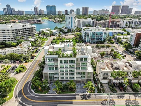 A modern building with a green roof is surrounded by other buildings and palm trees.