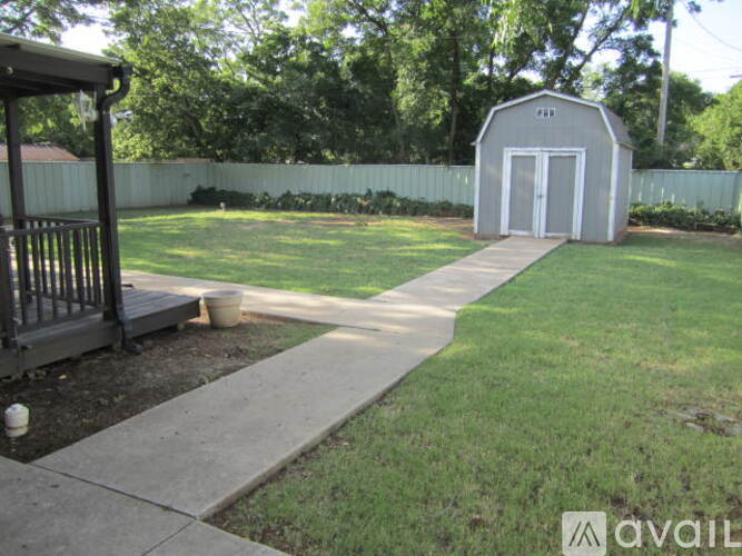 A small building with a white door is situated in a backyard with a concrete walkway leading to it.