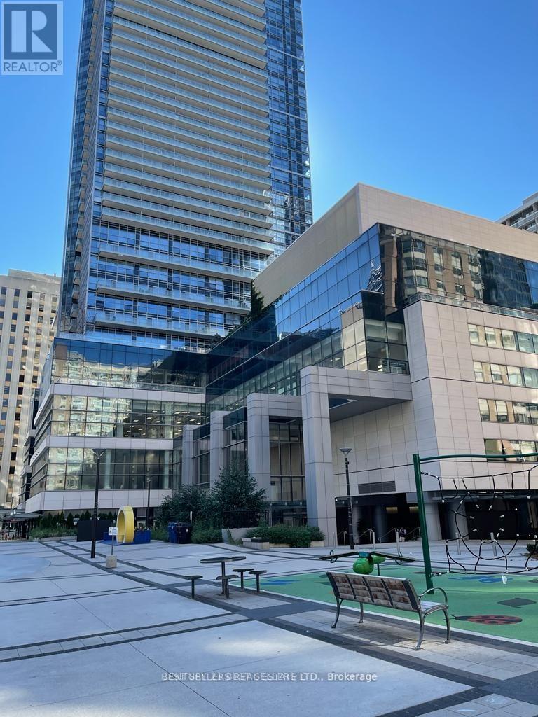 A large building with a glass facade is in the background of a park with a bench and a playground.