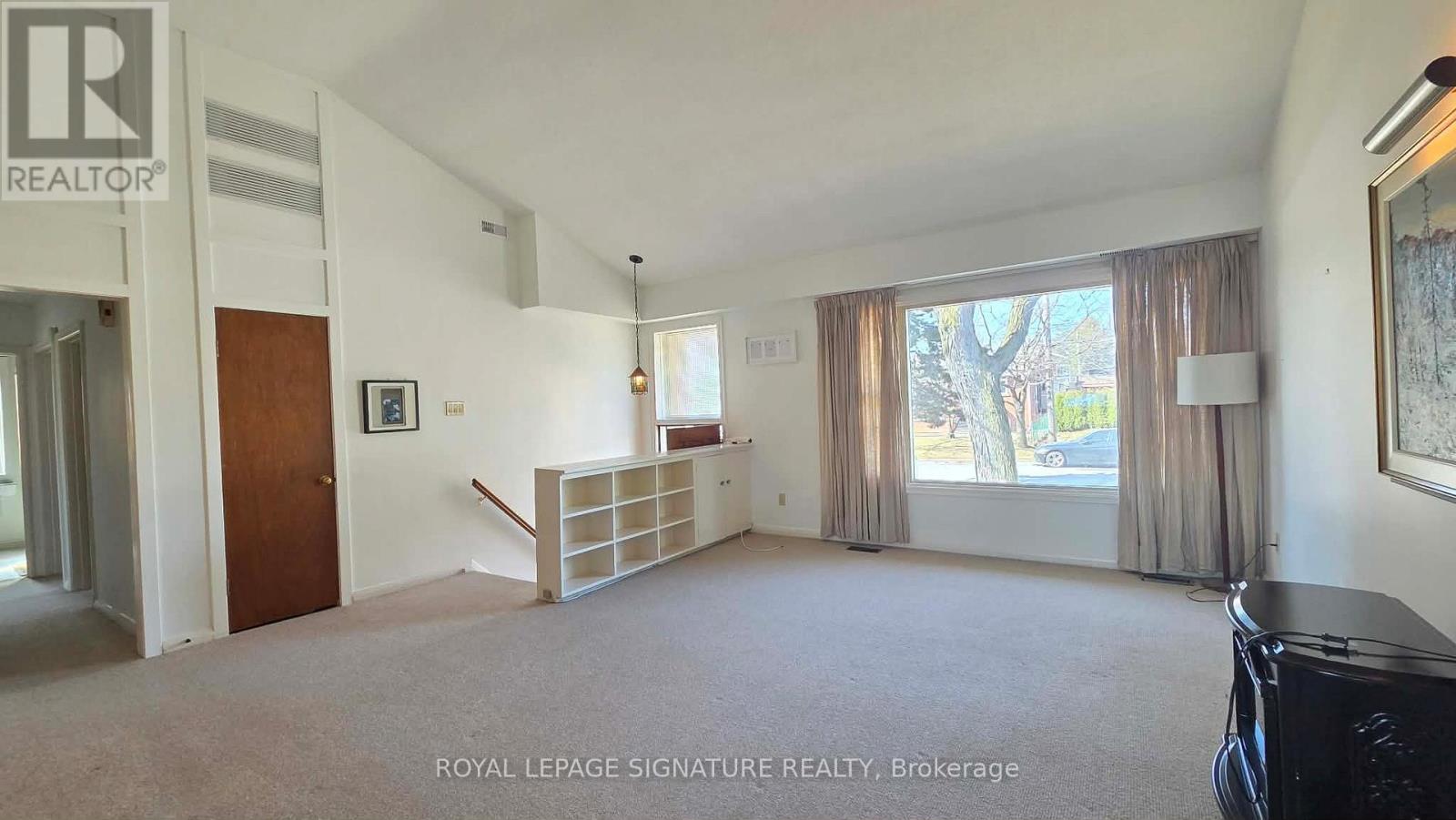 A spacious living room with a carpeted floor and a large window with curtains.