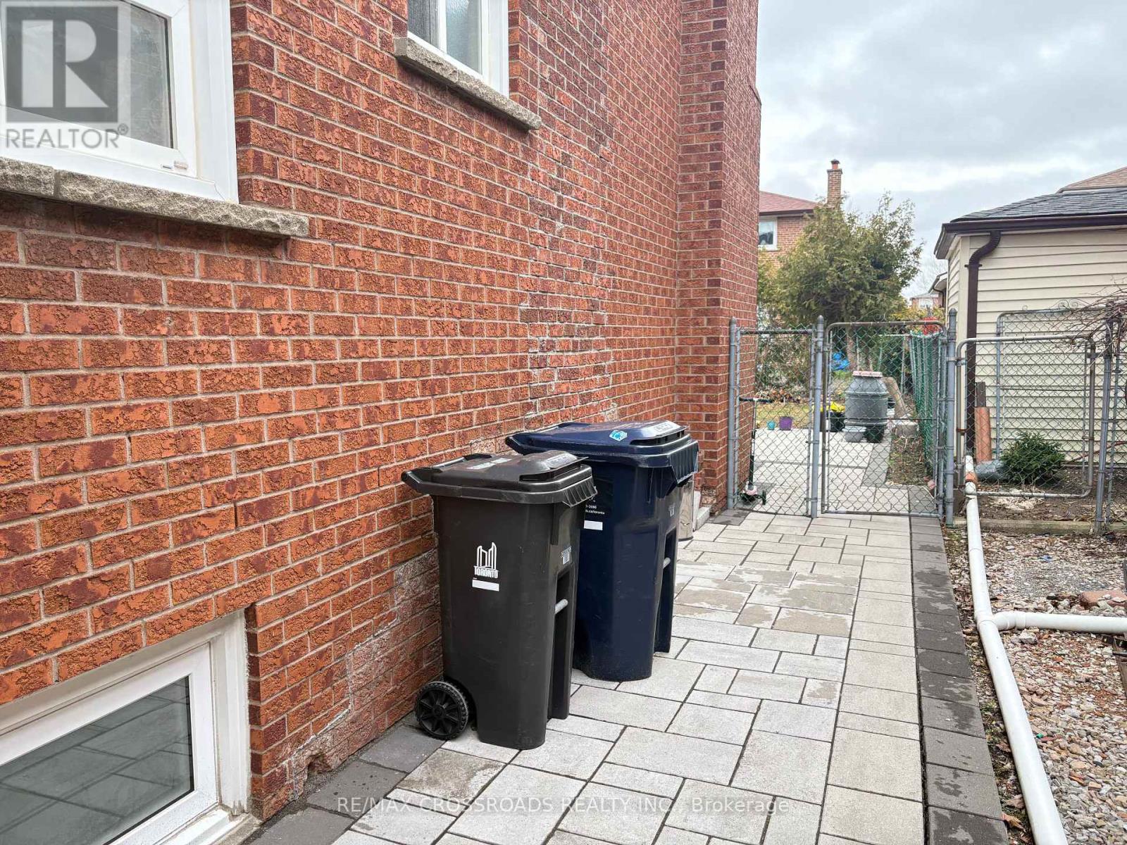 A black trash can with a white logo sits on a sidewalk next to a brick wall.