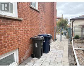 A black trash can with a white logo sits on a sidewalk next to a brick wall.