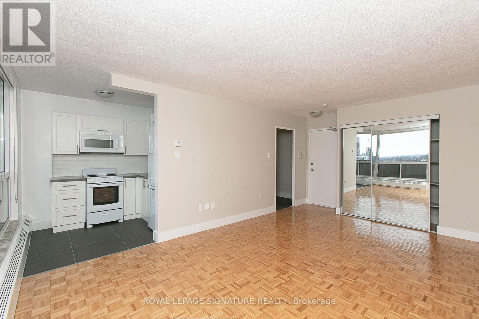 A spacious room with wooden flooring and a kitchen area with white appliances.