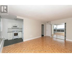 A spacious room with wooden flooring and a kitchen area with white appliances.