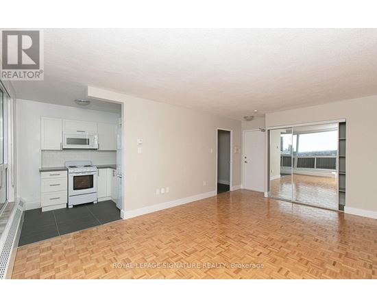 A spacious room with wooden flooring and a kitchen area with white appliances.