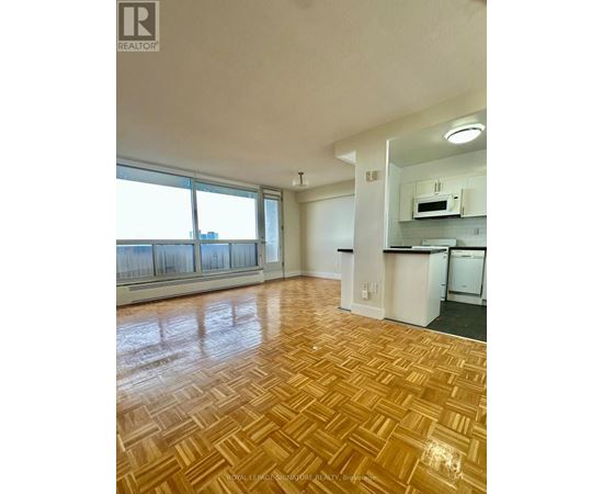 A room with wooden flooring and a kitchen area in the background.